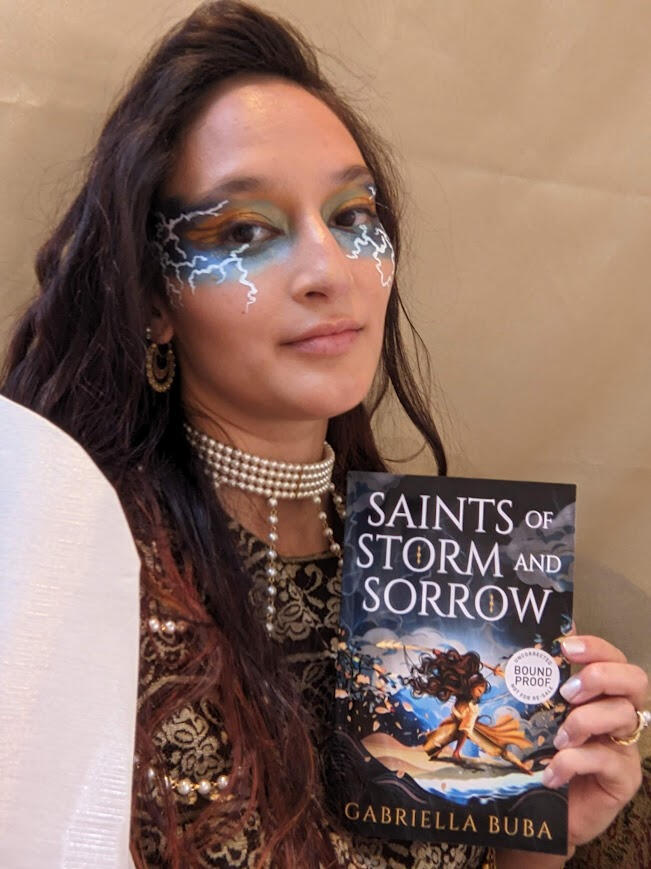 The author wearing gold and blue makeup that mimics the lightning on the cover of the book she's holding Saints of Storm and Sorrow. she is dark haired wearing white filipiniana sleeves and pearls