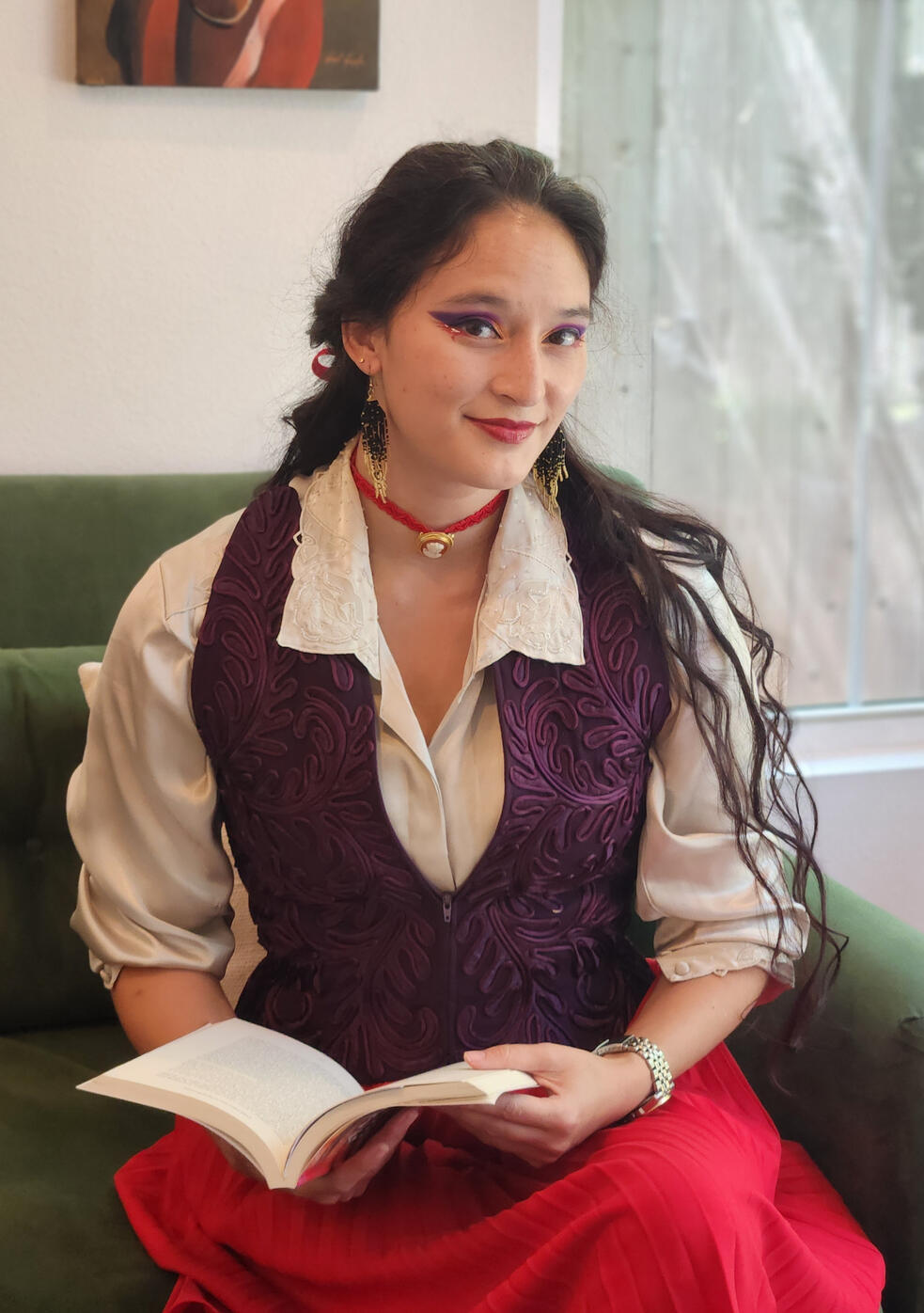 Headshot of the Author, long wavy dark haired woman wearing a collared shirt and purple vest, holding a book on a green couch
